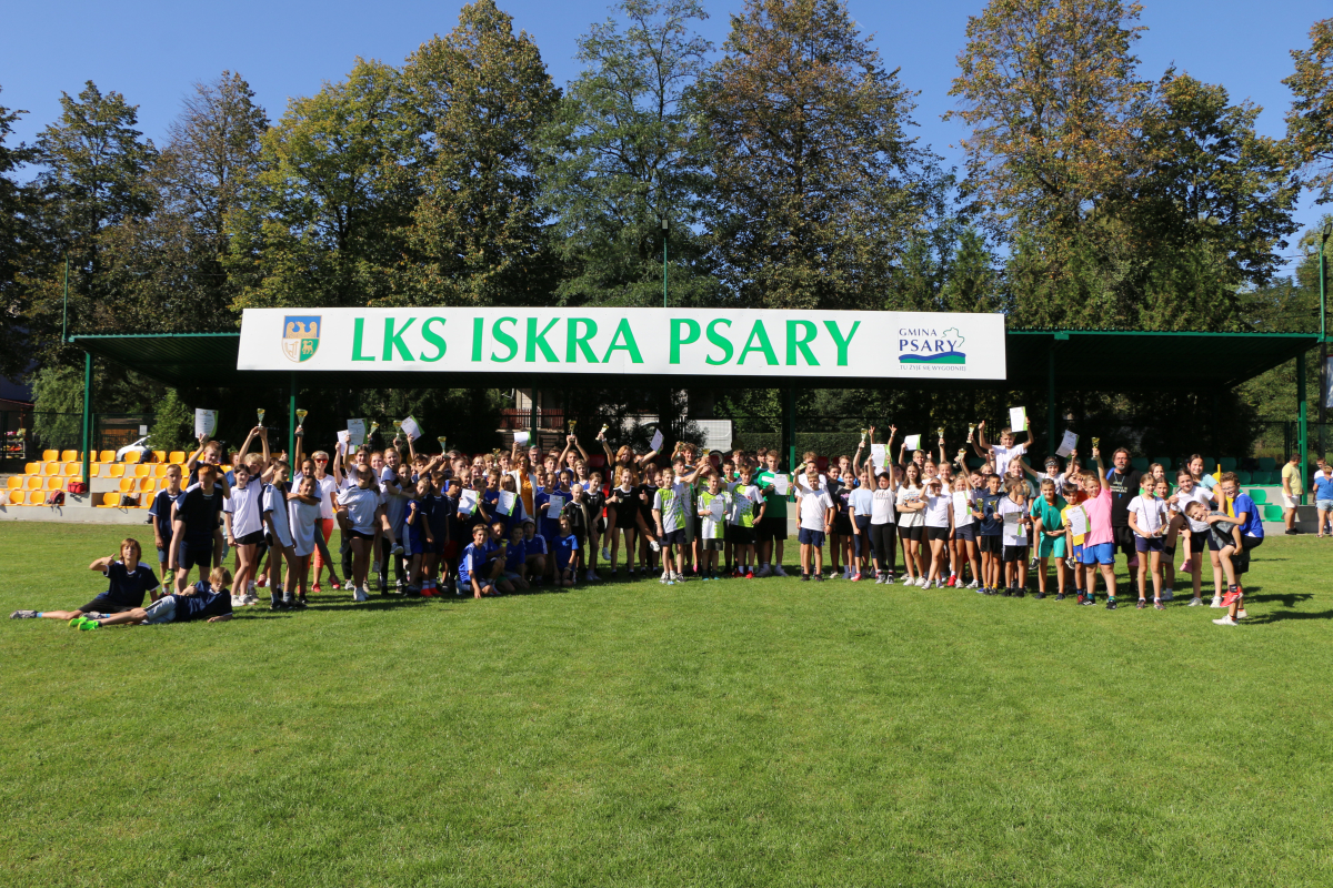 Zdjęcie przedstawia uczestników biegów sztafetowych z dyplomami i pucharami. W tle herb gminy, napis "LKS Iskra Psary" i logo gminy Psary. 