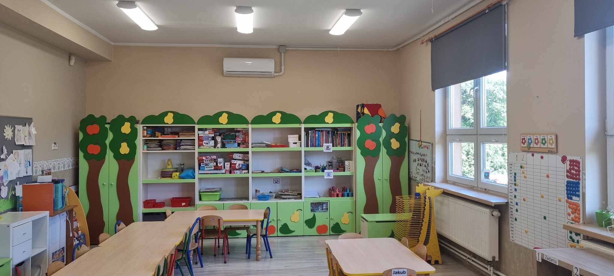 Na zdjęciu widać klasę przedszkolą z ławkami i krzesłami, kolorową ( z przewagą koloru zielonego) szafką na zabawki maluchów. W górnej części fotografii widać białą klimatyzację,a po prawej stronie - okna z ciemną roletą. 