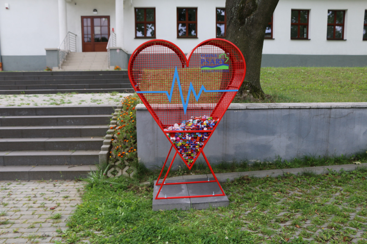 Na zdjęciu znajduje się duży, czerwony pojemnik w kształcie serca z logo Gminy Psary. W tle widać Ośrodek Kultury w Goląszy Górnej.