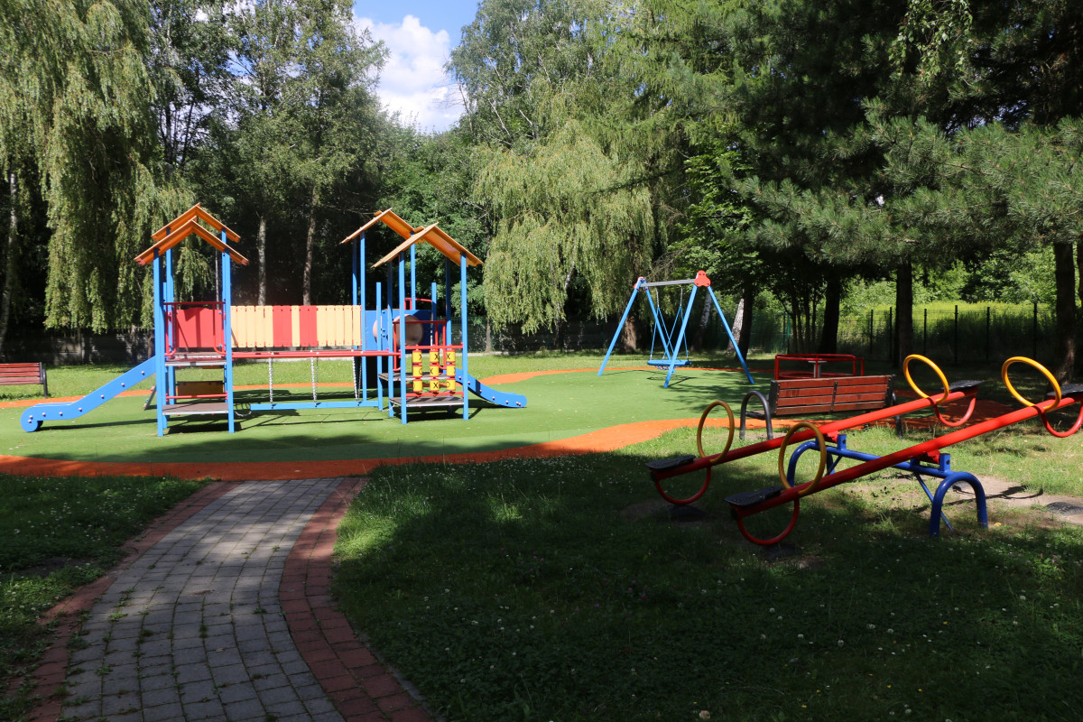 Zdjęcie przedstawia plac zabaw w przedszkolu w Sarnowie. Widzimy domek ze zjeżdżalną,huśtawkę z bocianim gniazdem i inne zabawki dla dzieci.