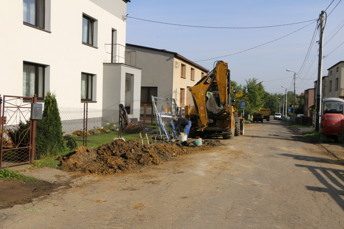 Zdjęcie przedstawia prace nad budową wodociągu. W centrum znajduje się koparka, pracownik oraz ziemia. Z Lewej strony znajdują się domy mieszkalne.