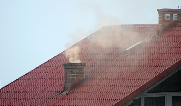 Zdjęcie przedstawia kawałek dachu z kominem, z którego wydobywa się dym. W tke niebieskie niebo