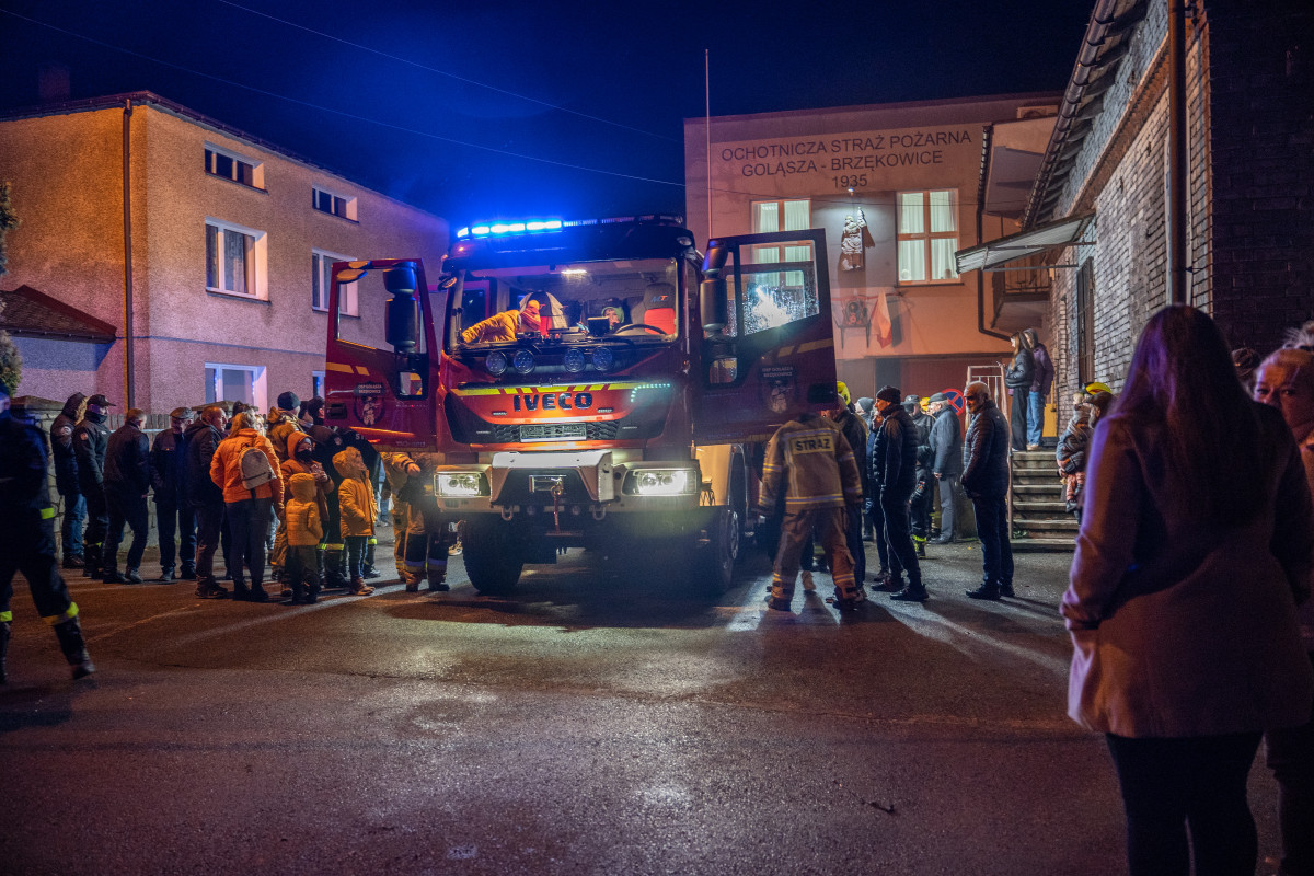 Fotografia przedstawia średni wóz ratowniczo-gaśniczy, wokół ludzie. Jest ciemno, pora wieczorowa