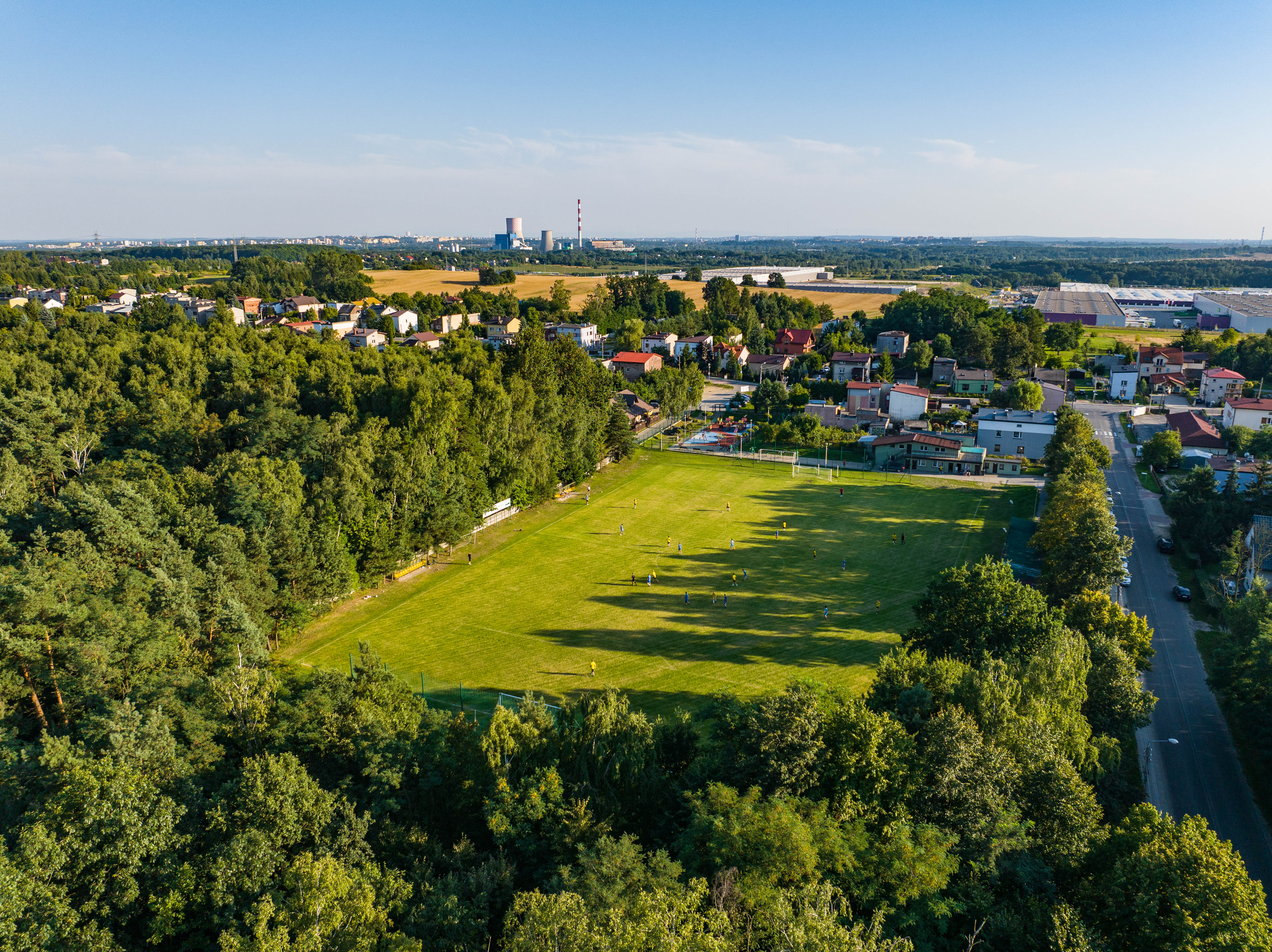 Zdjęcie przedstawia boisko sportowe z zawodnikami z lotu ptaka, dookoła las i panorama pobliskich miejscowości